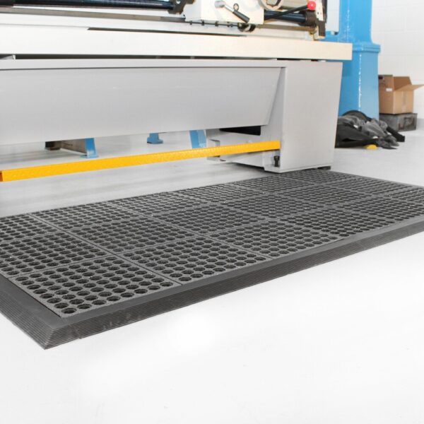 A Nitrile Top Interlocking Mat with holes in black is placed on a white floor in front of industrial machinery, with tools and boxes visible in the background.