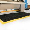 A Drainage Top Interlocking Mat with black anti-fatigue surface and yellow borders is positioned on a white floor in front of industrial machinery, with cardboard boxes and equipment visible in the background.