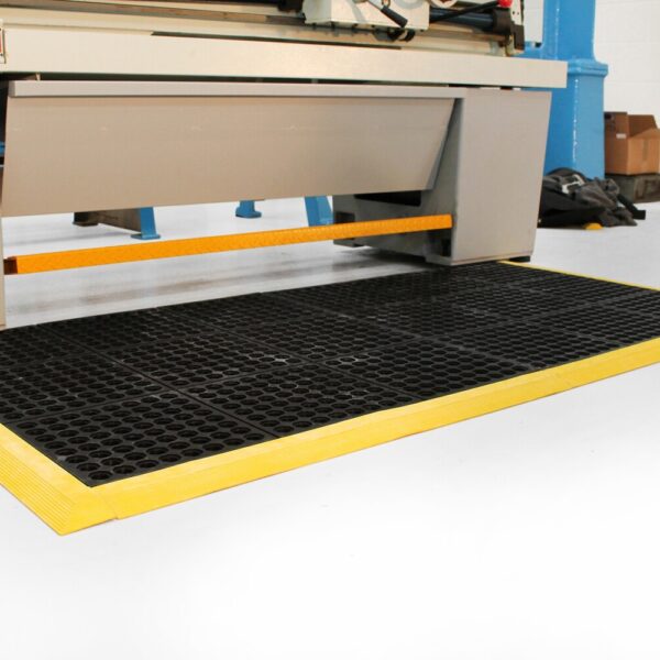 A Drainage Top Interlocking Mat with black anti-fatigue surface and yellow borders is positioned on a white floor in front of industrial machinery, with cardboard boxes and equipment visible in the background.