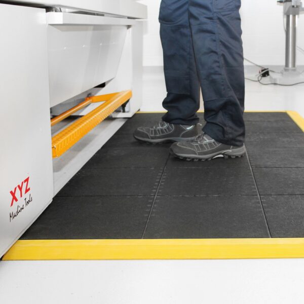 A person in dark trousers and shoes stands on a Solid Top Interlocking Mat with a yellow border beside an XYZ Machine Tools machine in a clean industrial setting.