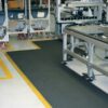 A factory or warehouse floor with Solid Top Interlocking Mats in black, yellow safety lines, mobile trolleys with red wheels, and metal workstations along a production or assembly line.