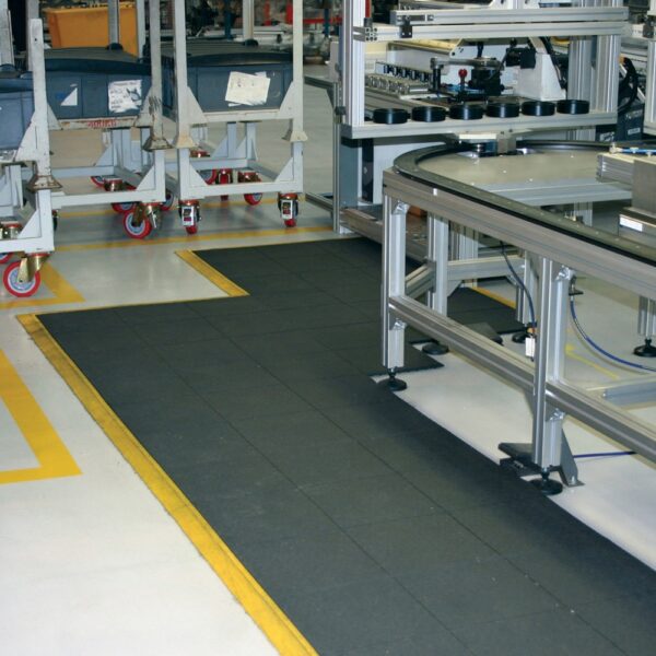 A factory or warehouse floor with Solid Top Interlocking Mats in black, yellow safety lines, mobile trolleys with red wheels, and metal workstations along a production or assembly line.