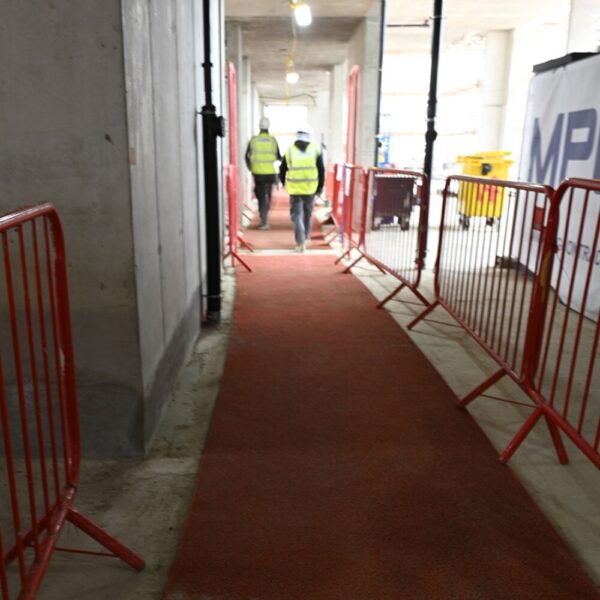 Two people in high-visibility vests walk down a path covered with heavy-duty PVC vinyl roll and lined by red barriers inside an unfinished construction building.