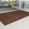 A Ribbed Doormat in brown rests on the tiled floor near the entrance of a building with glass doors, offering a view of cars parked outside.