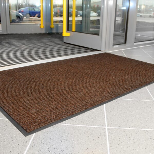 A Ribbed Doormat in brown rests on the tiled floor near the entrance of a building with glass doors, offering a view of cars parked outside.