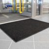 A Ribbed Doormat in black is placed on a light grey tiled floor inside a building entrance with glass doors featuring yellow handles, whilst cars can be seen parked outside.