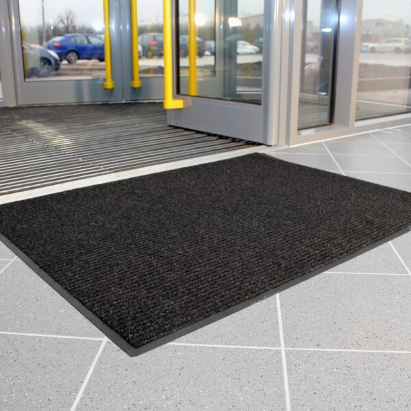 A Ribbed Doormat in black is placed on a light grey tiled floor inside a building entrance with glass doors featuring yellow handles, whilst cars can be seen parked outside.