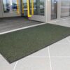 A Ribbed Doormat in dark green lies on a light grey tiled floor before glass entrance doors with yellow handles.
