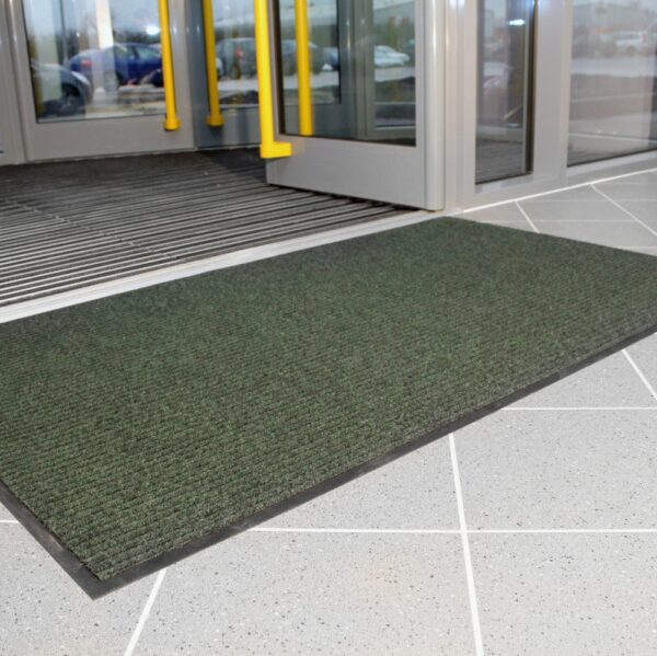 A Ribbed Doormat in dark green lies on a light grey tiled floor before glass entrance doors with yellow handles.