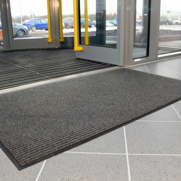 A Ribbed Doormat in grey is placed on a tiled floor in front of a glass entrance door, with parked cars visible outside through the windows.