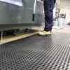 A person stands on a Drainage Workplace Mat beside industrial equipment; only their legs and beige shoes are visible. The mat’s textured surface and the clean, organised workspace highlight its suitability for industrial settings.