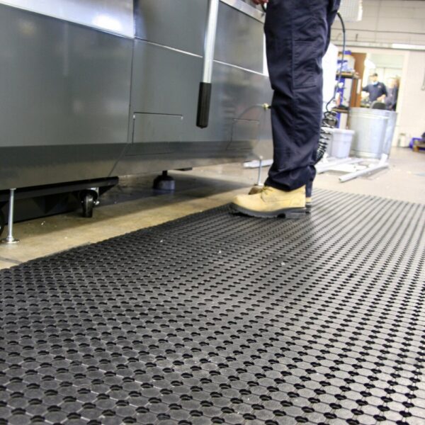A person stands on a Drainage Workplace Mat beside industrial equipment; only their legs and beige shoes are visible. The mat’s textured surface and the clean, organised workspace highlight its suitability for industrial settings.
