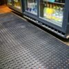 A Drainage Workplace Mat is placed on the floor in front of refrigerated glass display cases stocked with bottled fizzy drinks, water, and juices.