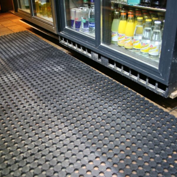 A Drainage Workplace Mat is placed on the floor in front of refrigerated glass display cases stocked with bottled fizzy drinks, water, and juices.