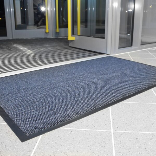 A Hard-Wearing Multi-Purpose Mat with a blue rectangular design and black border is on a light grey tiled floor in front of glass entrance automatic sliding doors.