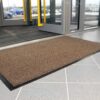 A Hard-Wearing Multi-Purpose Mat in brown sits on a grey tiled floor near glass entrance doors, with several cars visible outside through the windows.