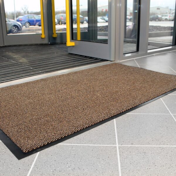 A Hard-Wearing Multi-Purpose Mat in brown sits on a grey tiled floor near glass entrance doors, with several cars visible outside through the windows.