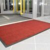 A Hard-Wearing Multi-Purpose Mat in red with a black border is laid on light grey tiles by an open glass entrance door. Yellow door handles and indoor lighting can be seen inside.