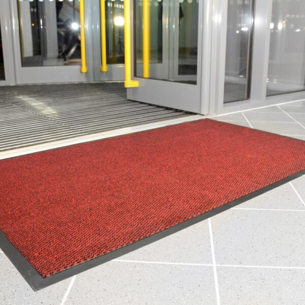 A Hard-Wearing Multi-Purpose Mat in red with a black border is laid on light grey tiles by an open glass entrance door. Yellow door handles and indoor lighting can be seen inside.