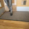 A person in grey suit trousers and black shoes steps onto the Hard-Wearing Multi-Purpose Mat, a brown mat placed inside a doorway with wooden doors and a wooden floor.