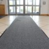 A Hard-Wearing Multi-Purpose Mat in grey runs along a tiled entryway towards glass double doors, with sunlight streaming in. A wooden cupboard and white walls are visible on the left.