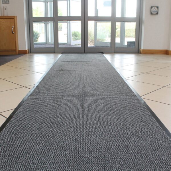 A Hard-Wearing Multi-Purpose Mat in grey runs along a tiled entryway towards glass double doors, with sunlight streaming in. A wooden cupboard and white walls are visible on the left.