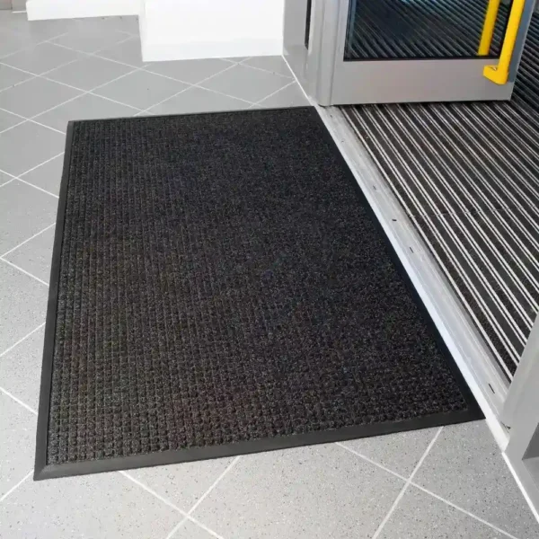 The Heavy Traffic Doorway Mat, in black and rectangular shape, is placed on a grey tiled floor by the entrance of a building with the door open.