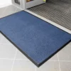 The Heavy Traffic Doorway Mat, featuring a blue textured surface and black border, is positioned on a light grey tiled floor near the building’s entrance with a metal threshold.