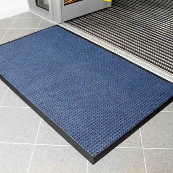 The Heavy Traffic Doorway Mat, featuring a blue textured surface and black border, is positioned on a light grey tiled floor near the building’s entrance with a metal threshold.