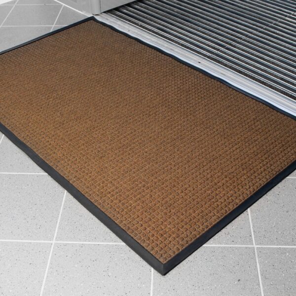 The Heavy Traffic Doorway Mat, featuring a brown textured surface and black border, is placed on a light grey tiled floor in front of a doorway with a metal threshold.