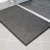The Heavy Traffic Doorway Mat, a dark grey rectangular mat with a textured surface, is placed on a light grey tiled floor near an open doorway with a metal grating just inside the entrance.