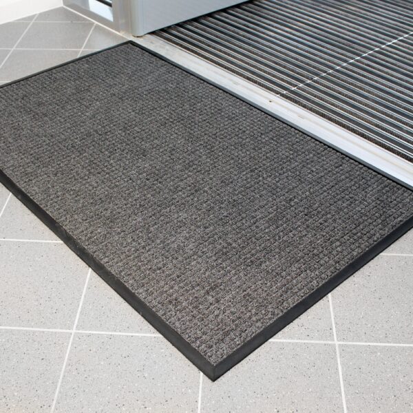The Heavy Traffic Doorway Mat, a dark grey rectangular mat with a textured surface, is placed on a light grey tiled floor near an open doorway with a metal grating just inside the entrance.