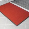 The Heavy Traffic Doorway Mat, featuring a red textured design with a black border, sits on a light grey tiled floor by a door with a metal threshold.