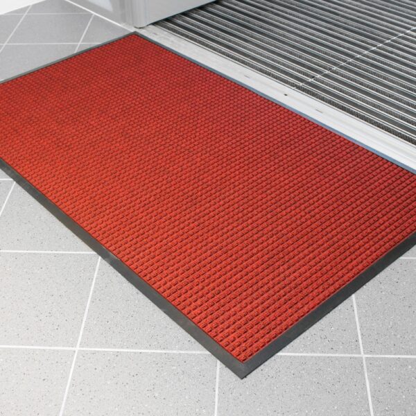 The Heavy Traffic Doorway Mat, featuring a red textured design with a black border, sits on a light grey tiled floor by a door with a metal threshold.