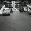 Close-up of a gym floor featuring interlocking gym mats with black rubber finish, various dumbbells scattered around, and racks of additional weights and gym equipment visible in the background.