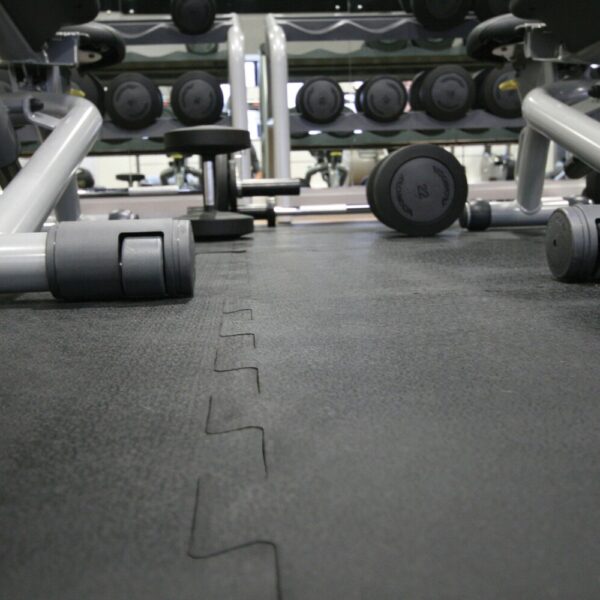 Close-up of a gym floor featuring interlocking gym mats with black rubber finish, various dumbbells scattered around, and racks of additional weights and gym equipment visible in the background.