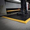 A person in black safety boots stands on a Heavy-Duty Workstation Mat with yellow edges in an industrial area near metal machinery.