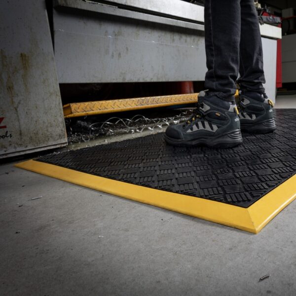 A person in black safety boots stands on a Heavy-Duty Workstation Mat with yellow edges in an industrial area near metal machinery.