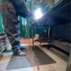 A person in protective clothing and boots is welding metal in a workshop, with bright sparks flying as they work on a Diamond Welding Mat.