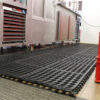 Close-up of the Drainage Top Interlocking Mat, a black rubber anti-fatigue mat with circular holes, shown on a kitchen or industrial floor in front of stainless steel appliances and cupboards.