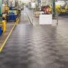 A clean, well-lit factory floor with industrial machines and safety barriers along a wide walkway fitted with Circular Stud Interlocking Tile, featuring a black surface and yellow dashed safety line for clear marking.