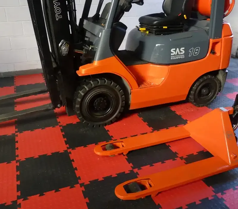 An orange forklift and an orange pallet truck are positioned on a red and black chequered floor with a white brick wall in the background.