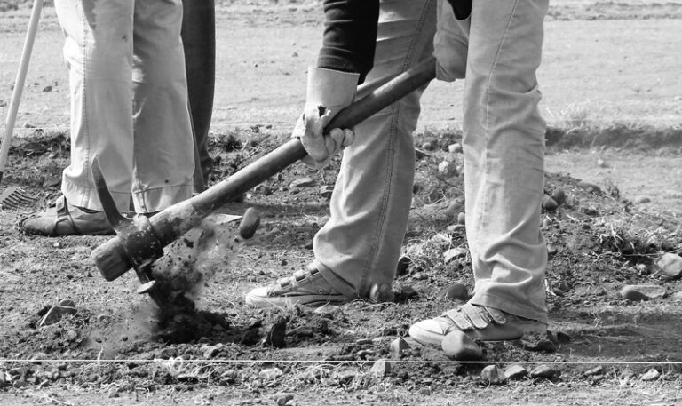 Two people working outdoors, using tools to dig and break up soil. One person wields a pickaxe, while the other stands nearby with a rake. The image is in black and white, focusing on their legs and hands.