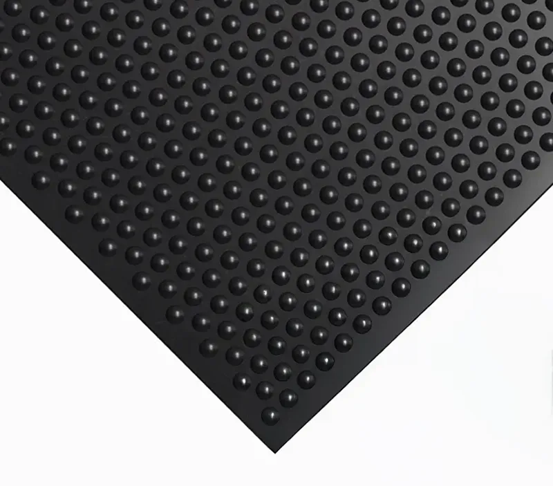 A black rubber mat with a pattern of raised, rounded bumps is shown on a white background. The image focuses on the corner of the mat, highlighting its textured surface.