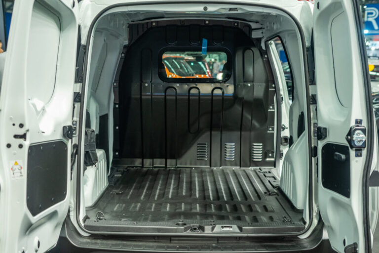 The image shows the empty cargo area of a white delivery van with both rear doors open, revealing a spacious, black-rubber-lined interior ready for transporting goods.