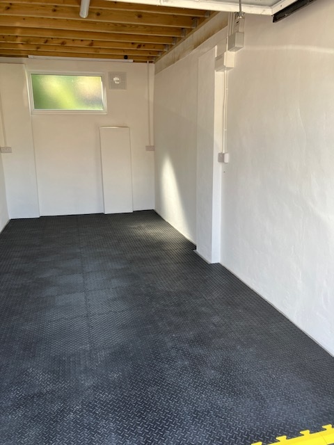 A clean, empty room with white walls, black textured flooring, an exposed wooden ceiling, and a frosted window letting in natural light.