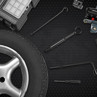 A car tyre, a hubcap, and various car repair tools, including a jack, screwdriver, spanner, and a wheel brace, are arranged on a black textured surface.