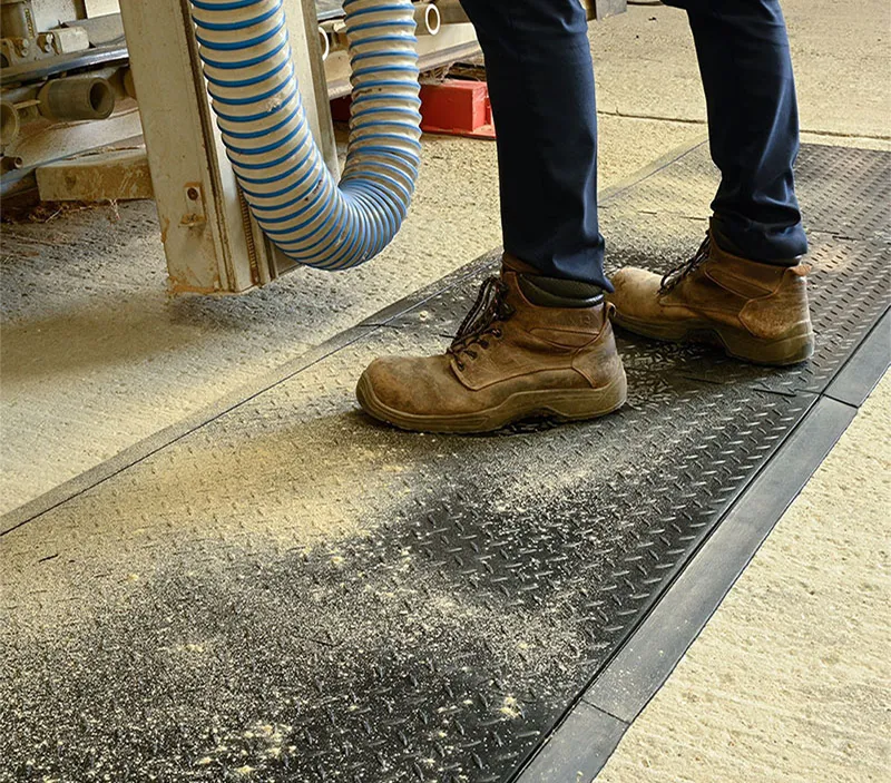 A person wearing brown work boots stands on a textured anti-fatigue mat with sawdust scattered around, next to machinery and a blue-striped hose in an industrial setting.