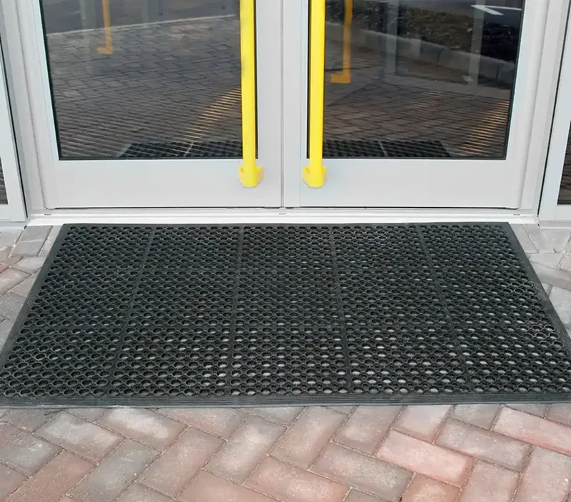 A black rubber doormat with circular holes is placed in front of a glass door with yellow handles. The area outside consists of interlocking stone paving.