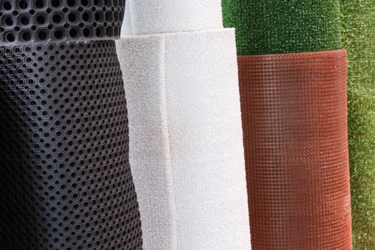 Five large rolls of different textured materials are standing vertically side by side: black with circular holes, white textured, white smooth, brown textured, and green artificial grass.
